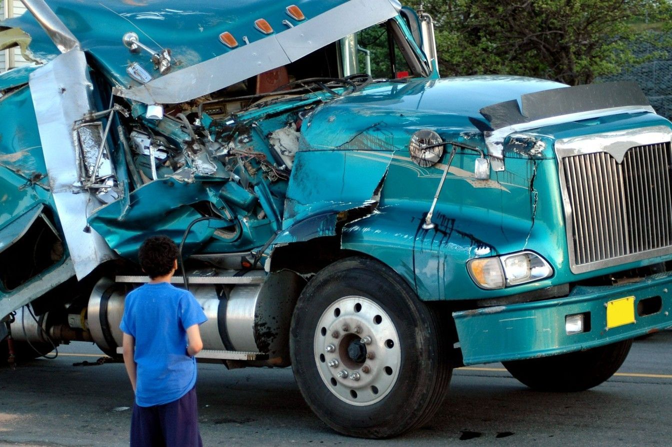 A boy in a blue shirt stands in front of a damaged semi truck