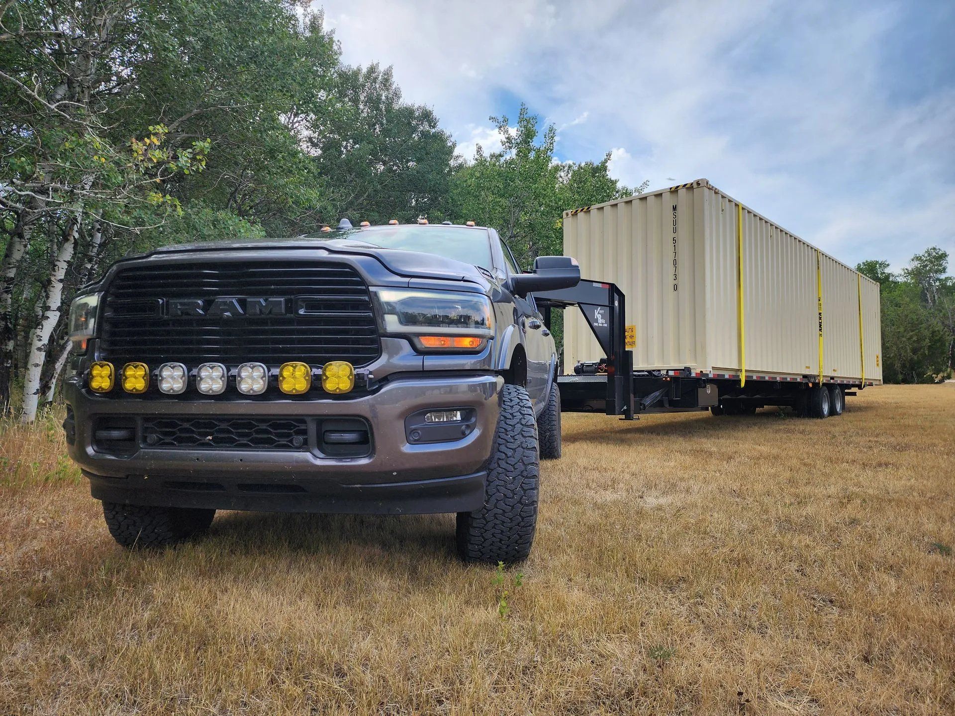 Brown RAM truck towing a shipping container on a flatbed trailer in a grassy field.