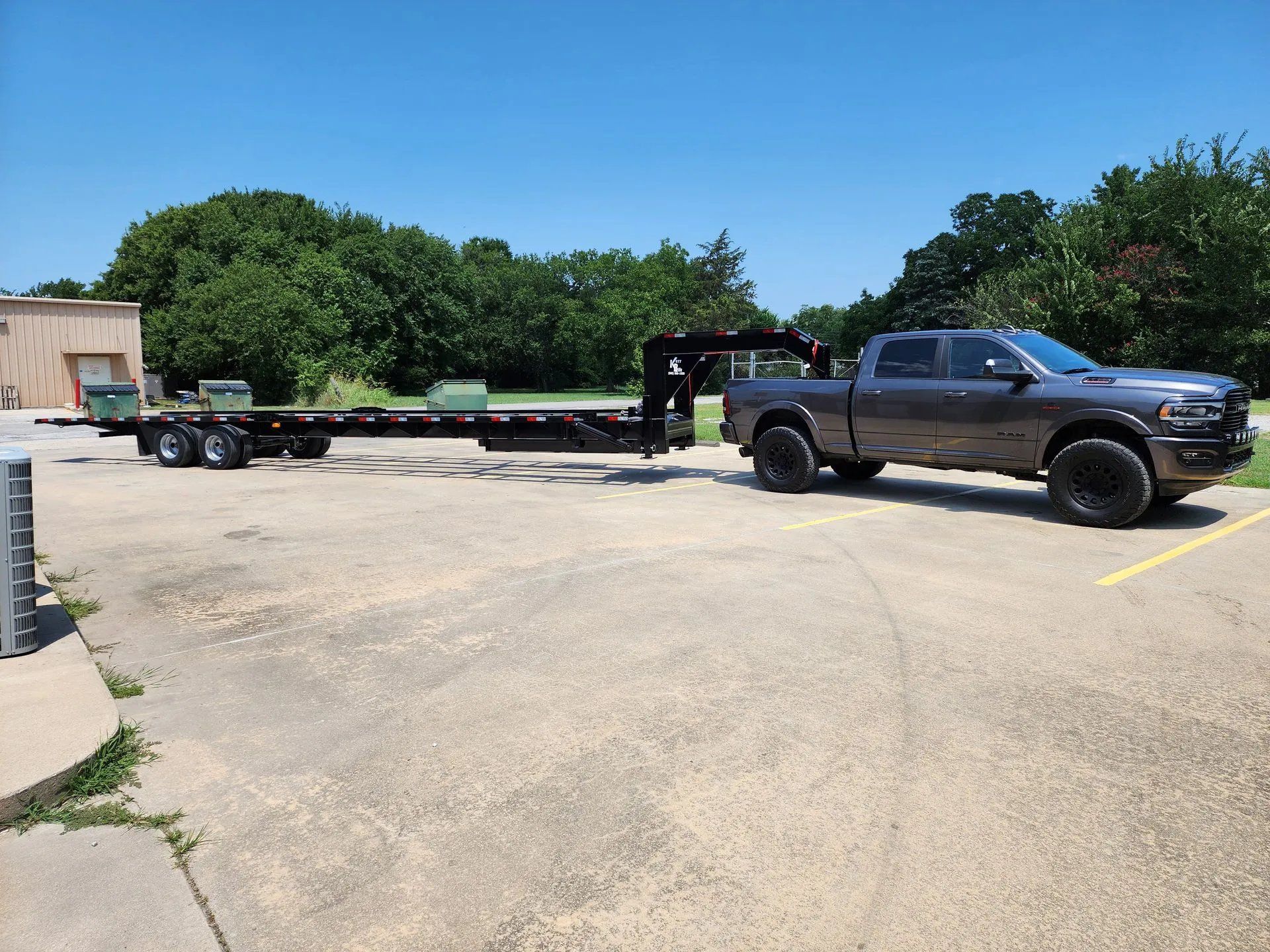 Gray pickup truck towing a long black trailer parked on a concrete surface. Green trees and a building in the background.
