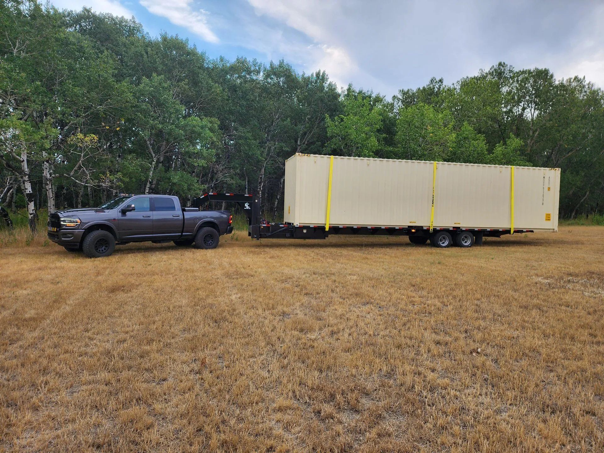 Dark gray truck towing a long beige container on a trailer in a grassy field with trees.