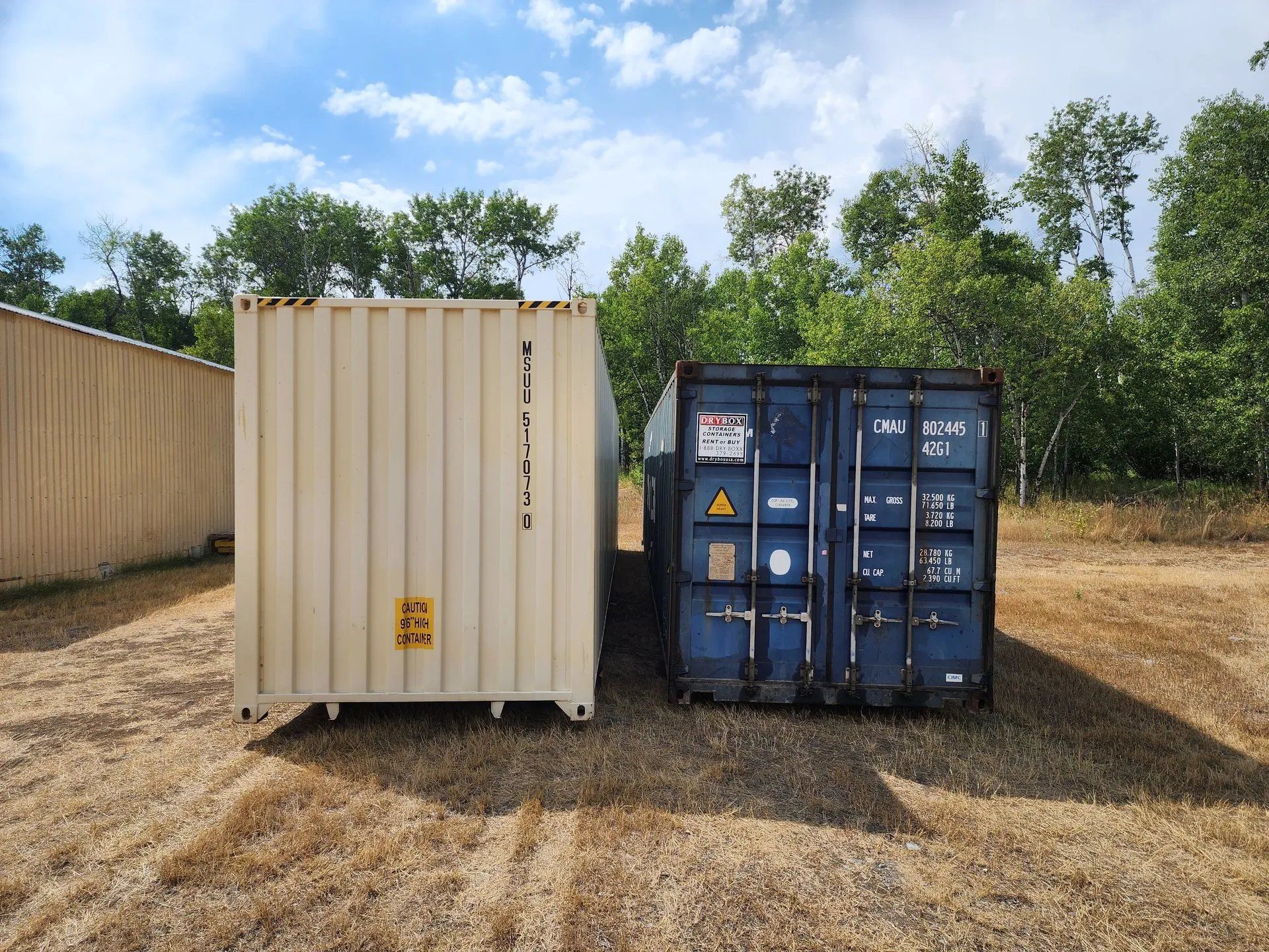 Two shipping containers, beige and blue, side-by-side on dry grass, with a building and trees in the background.
