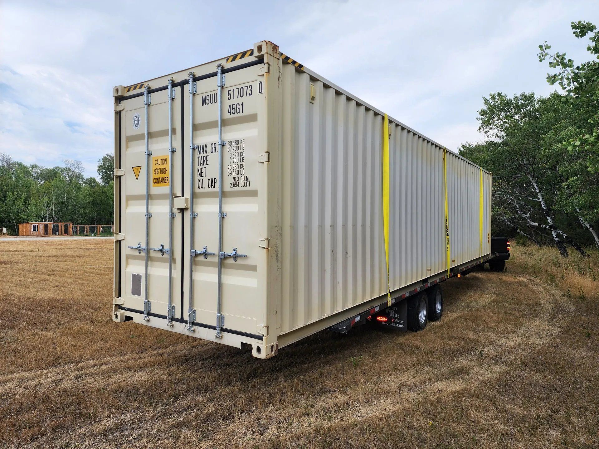 Beige shipping container on a trailer parked in a field with trees in the background.