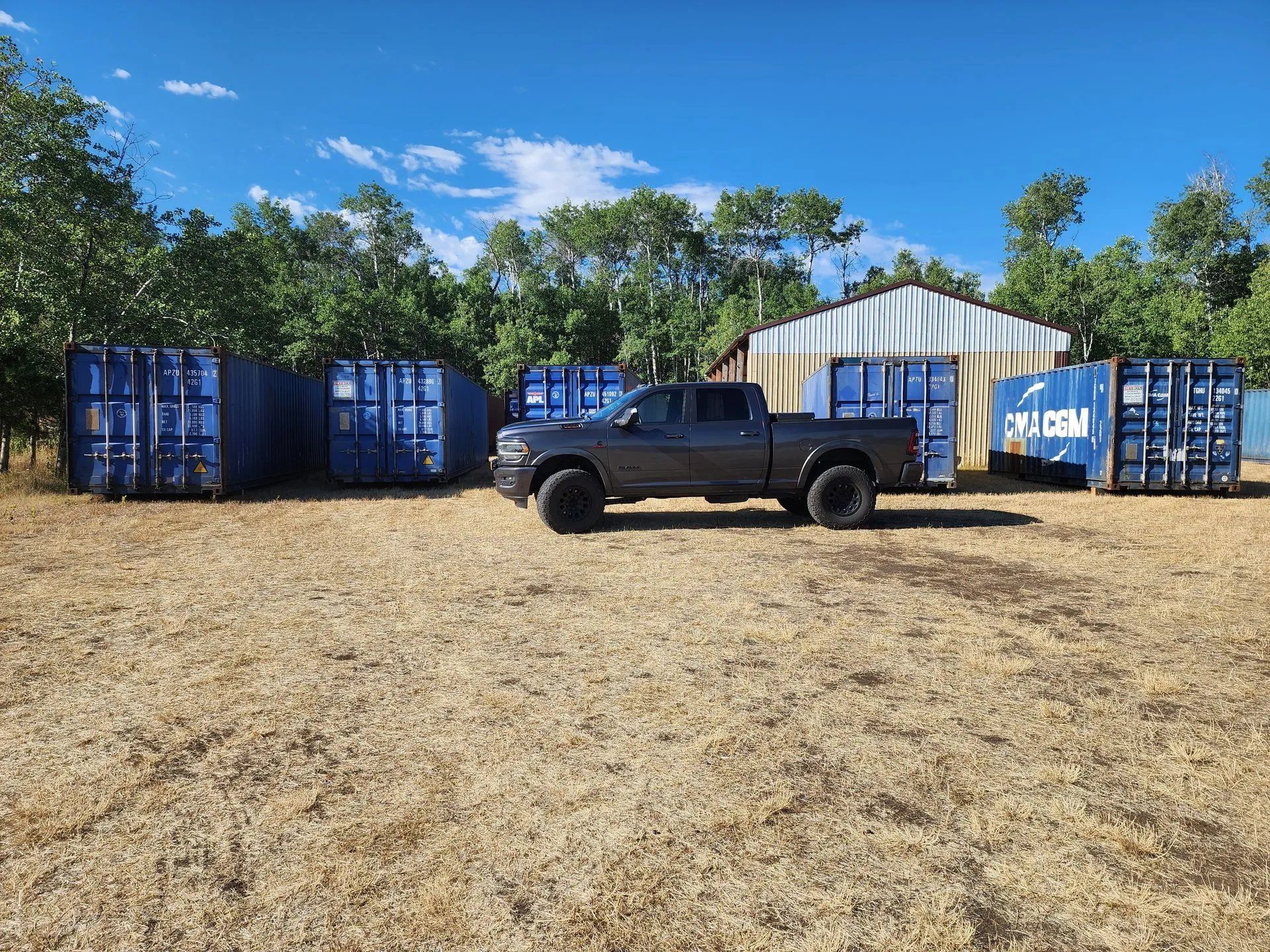 Gray truck parked near blue storage containers and a building on dry grass under a blue sky.