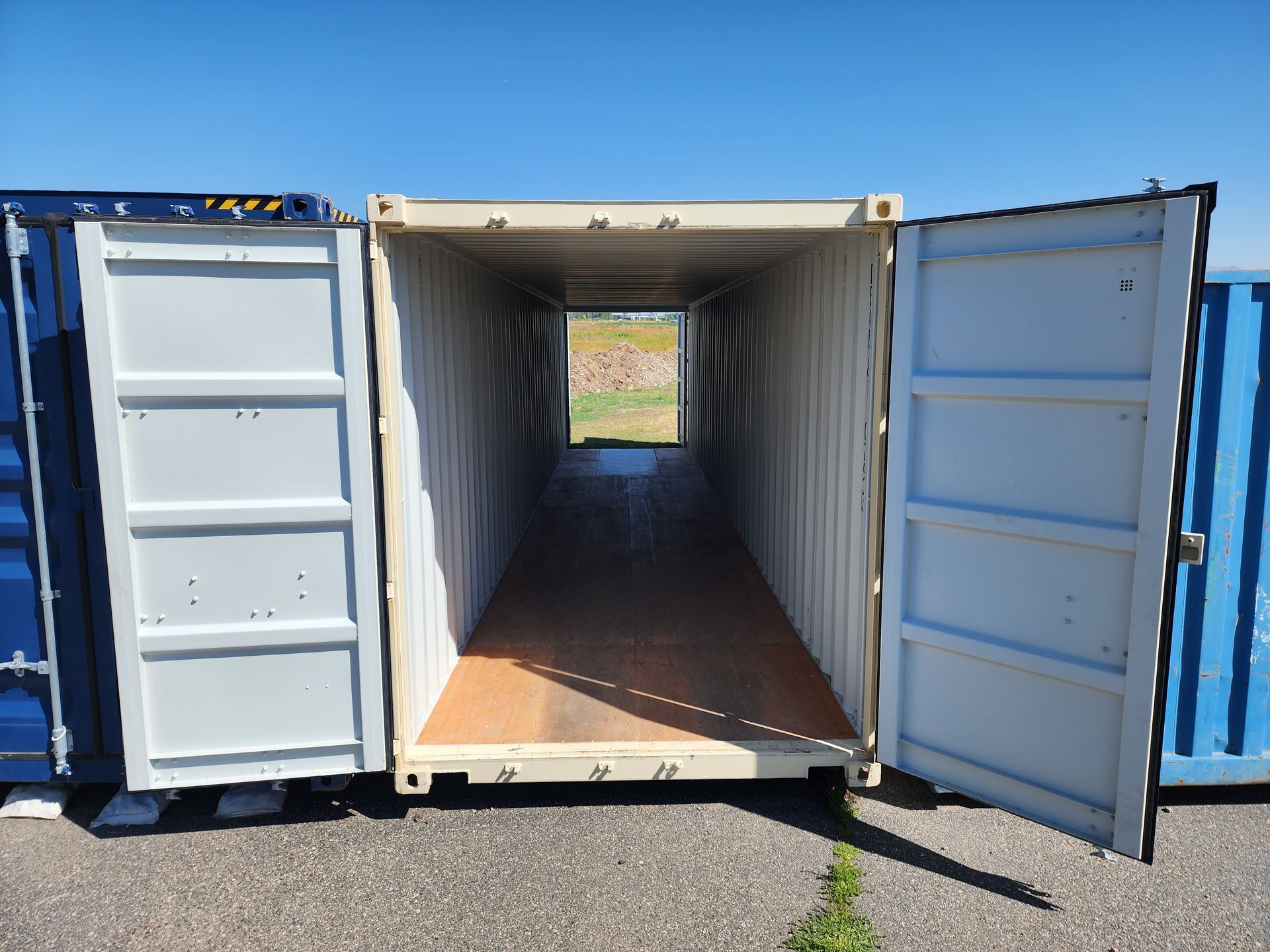 Open beige shipping container, doors ajar, revealing interior, with blue containers on either side, sunny outdoor setting.