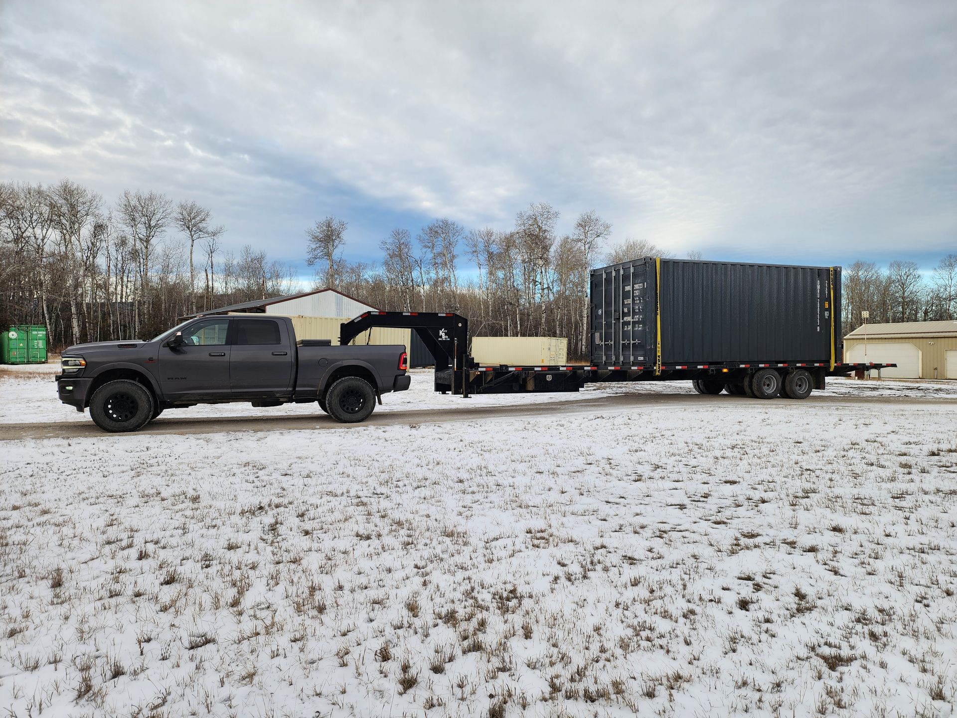 Gray truck towing a dark container on a trailer across a snow-covered field.