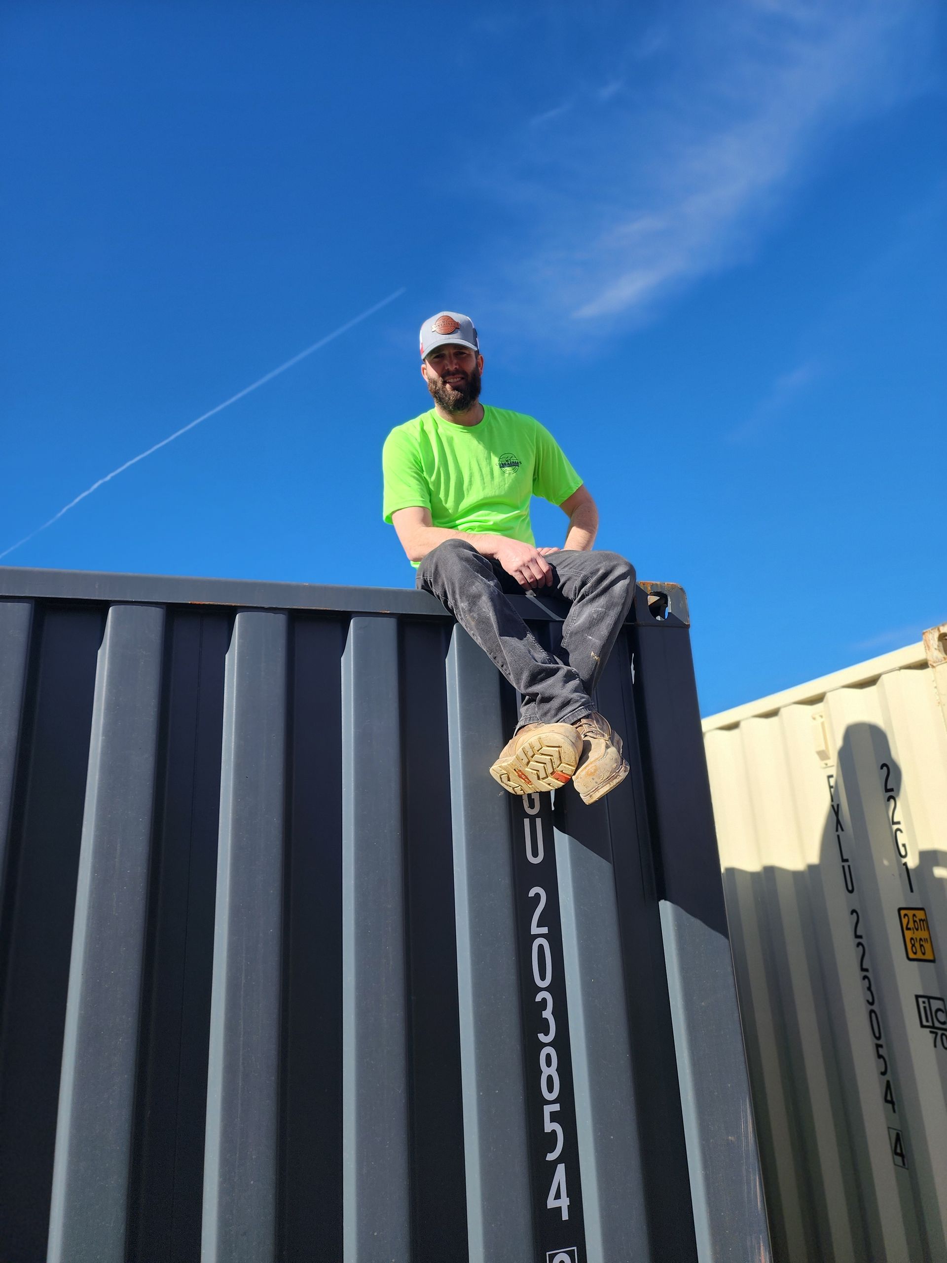 Man in green shirt sits atop a shipping container against a blue sky.