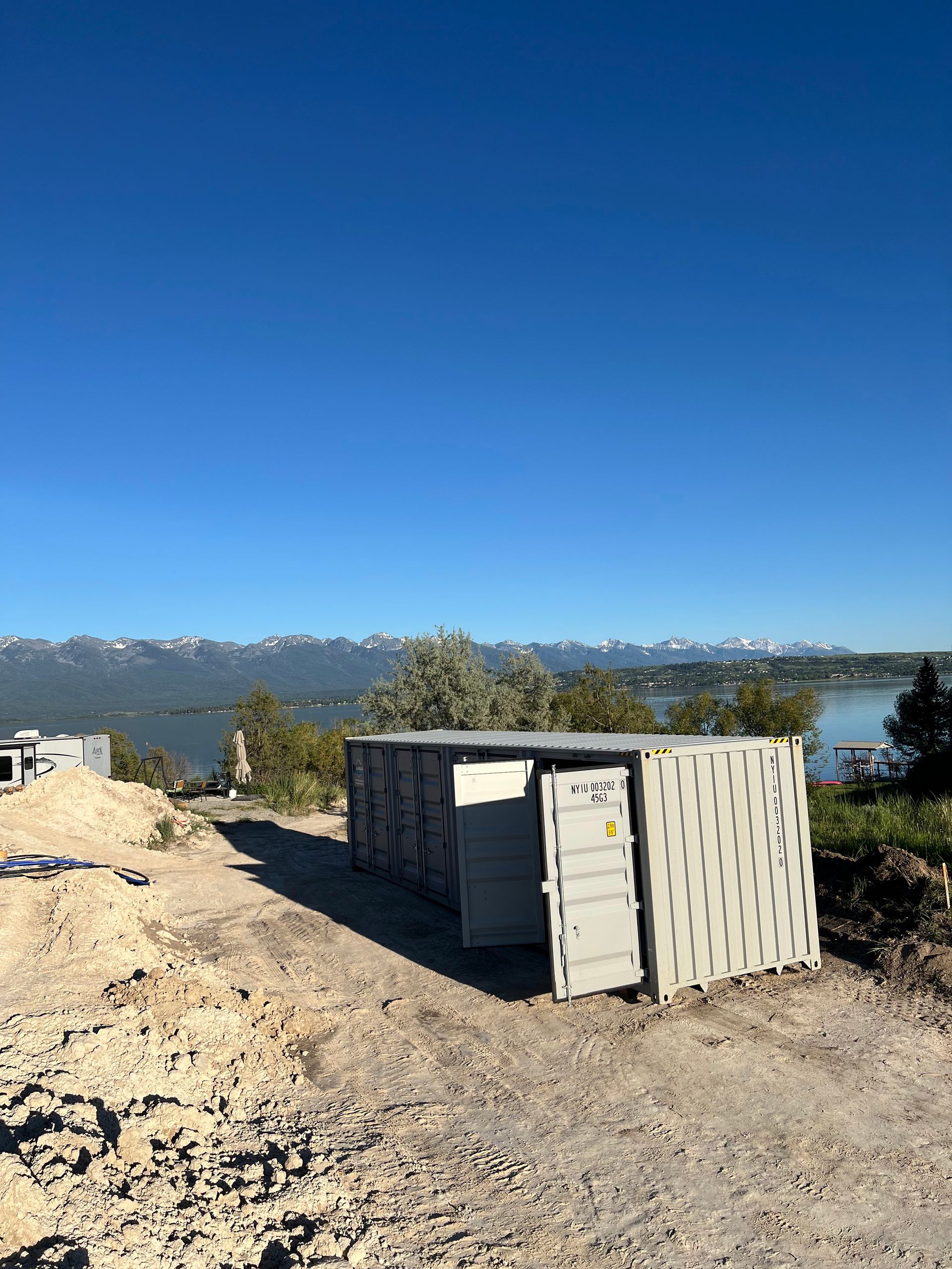 Gray container with open door on dirt path, overlooking water and mountains under a blue sky.