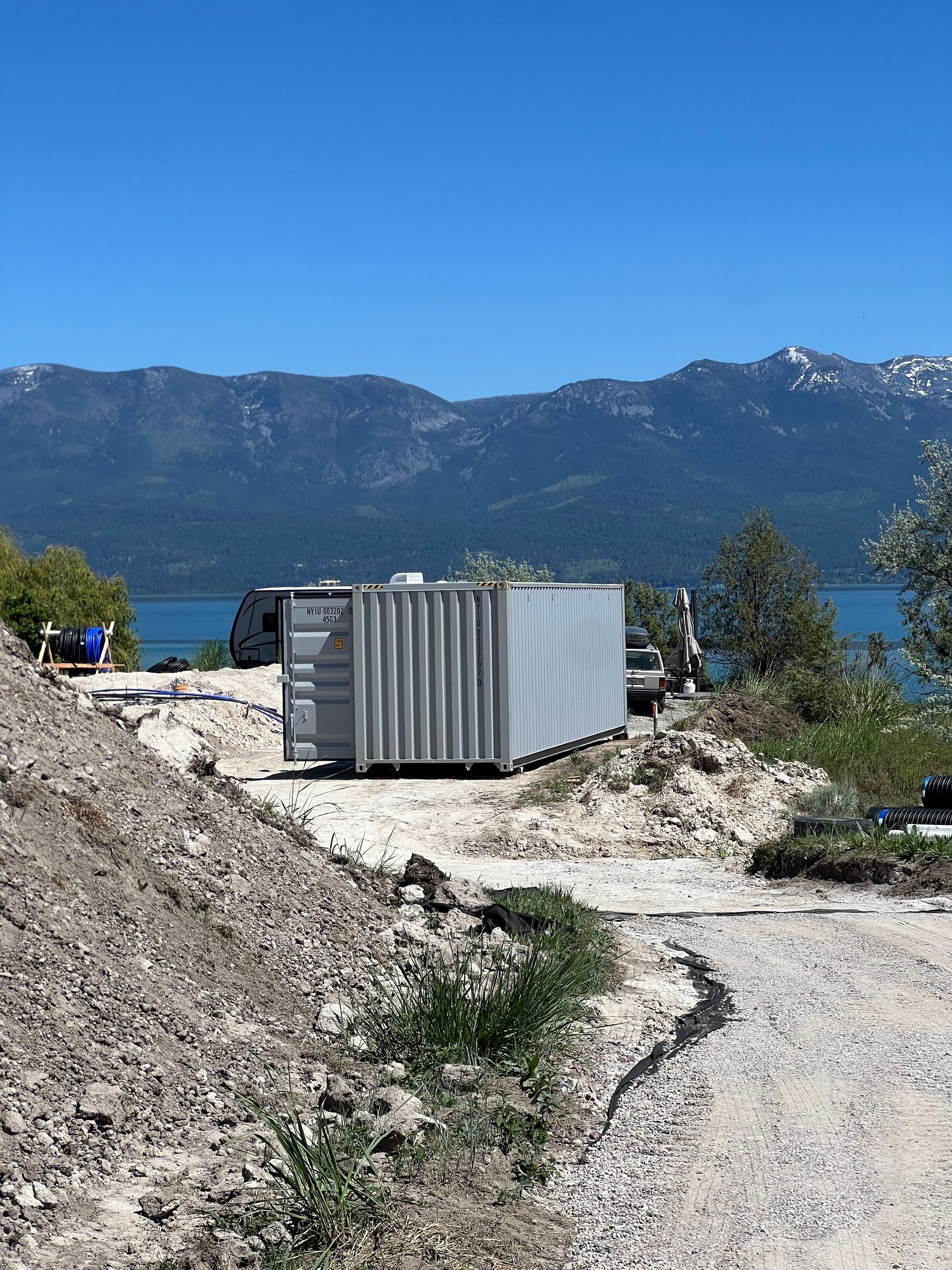Gray shipping container near water and mountains, under a blue sky.