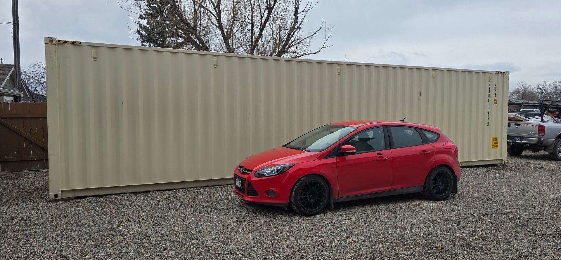 Red car parked near a large tan shipping container on a gravel surface. Cloudy day.