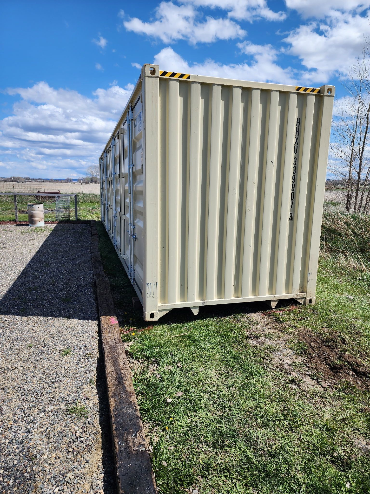Beige shipping container on gravel and grass under a blue sky.