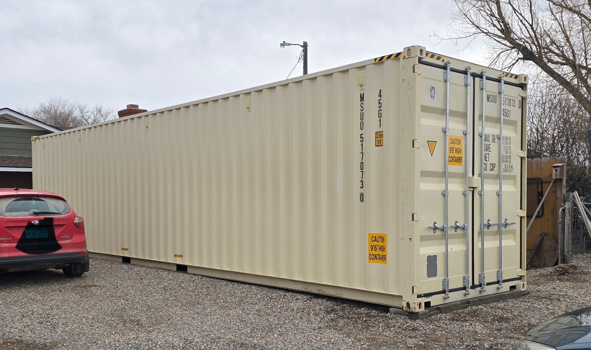 Tan shipping container in a gravel yard next to a red car and a building on a cloudy day.