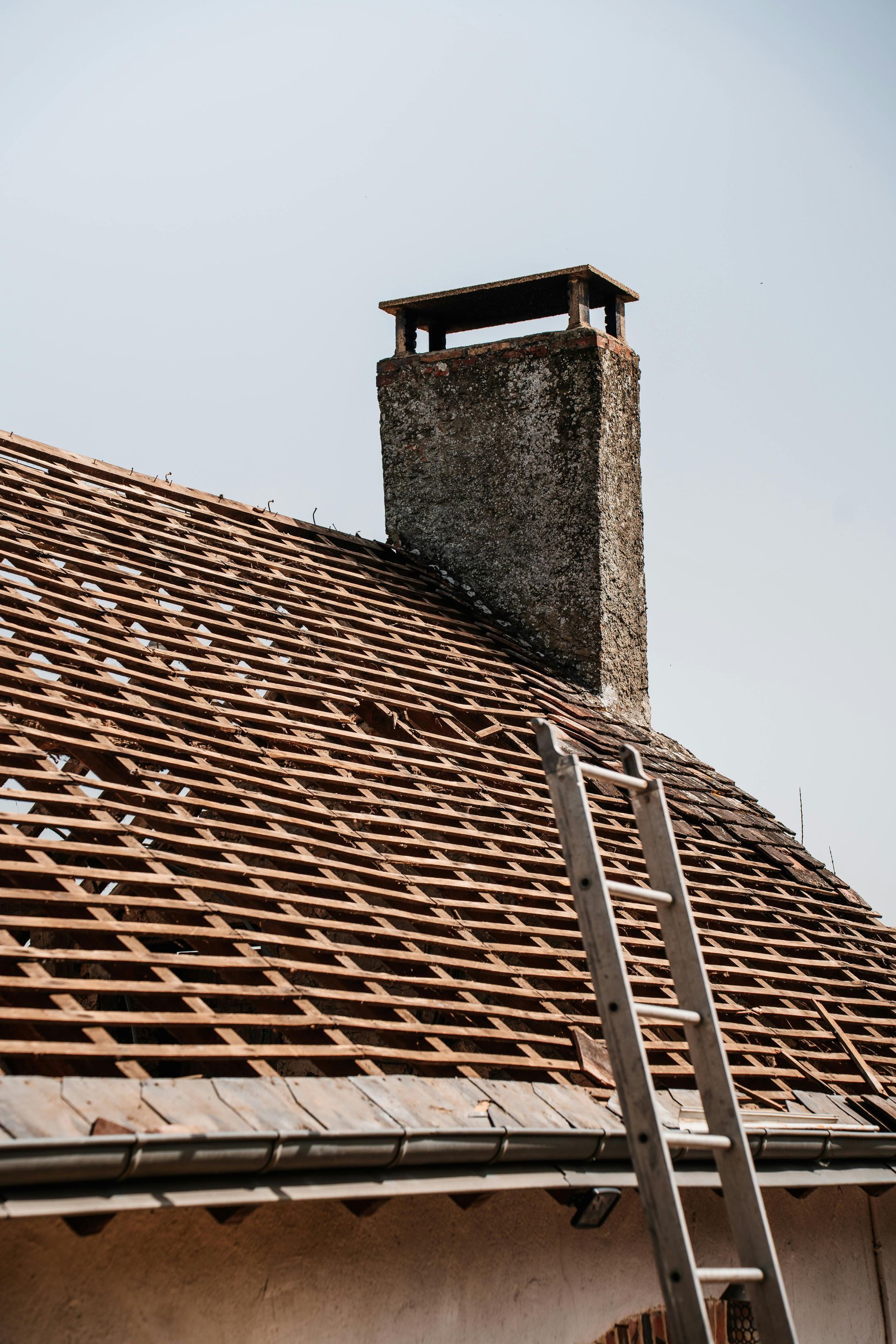 Roof with missing tiles, wooden slats exposed, tall chimney, ladder leaning against the roof.