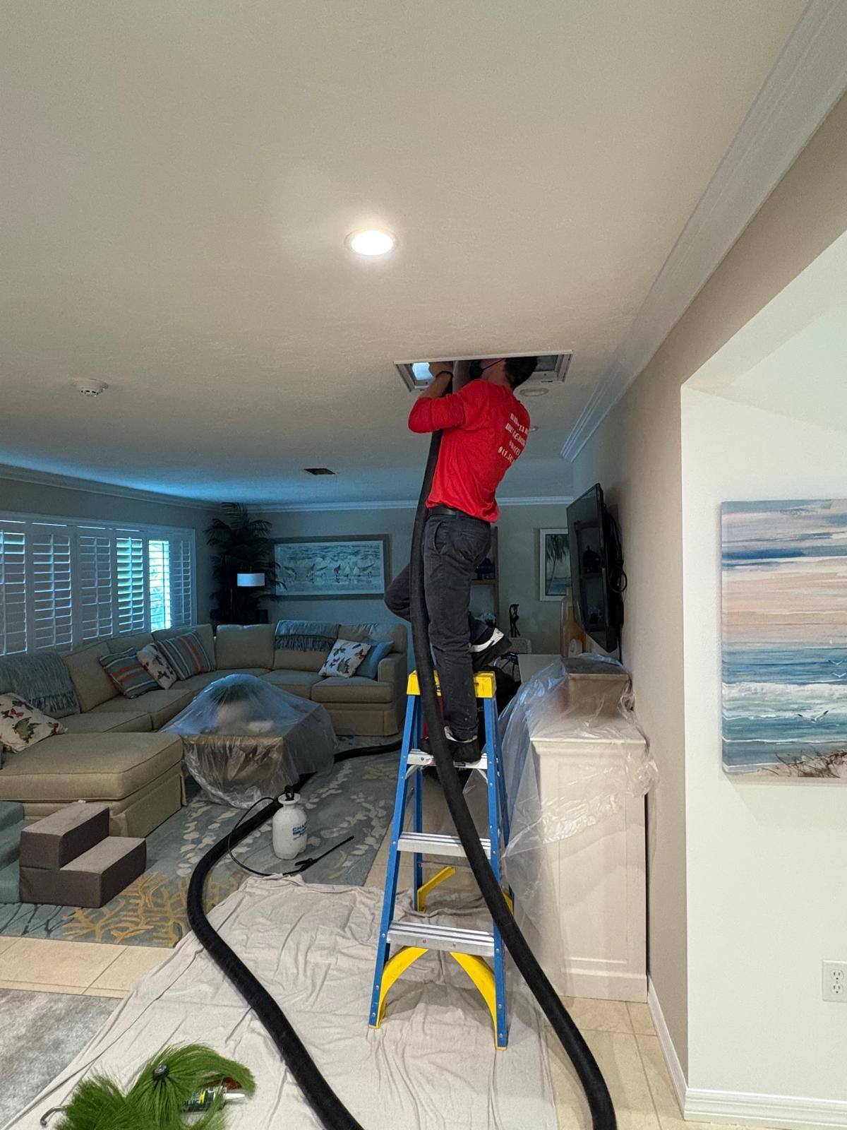 Person on ladder cleaning an air vent in a living room. Black hose extends down.