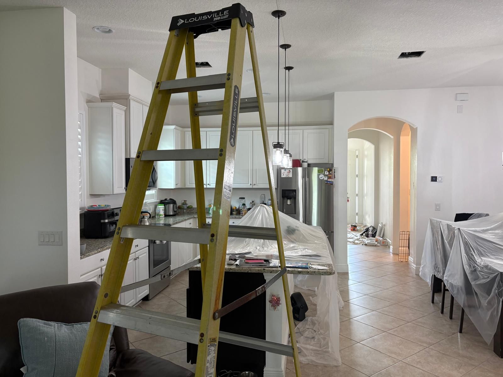 Yellow ladder in a kitchen, island covered in plastic sheeting. Kitchen cabinets, pendant lights visible.