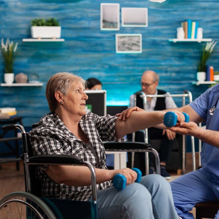 Une femme âgée en fauteuil roulant est aidée par une infirmière avec des haltères.