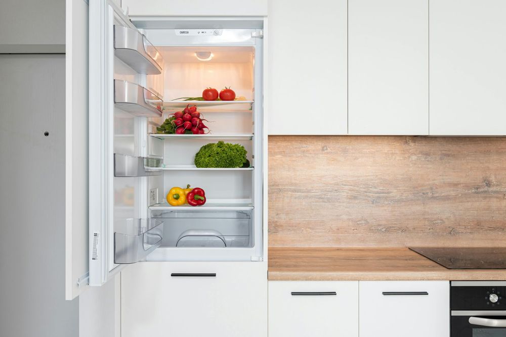 Open white refrigerator stocked with fresh vegetables in a modern, minimalist kitchen with light wood countertops.
