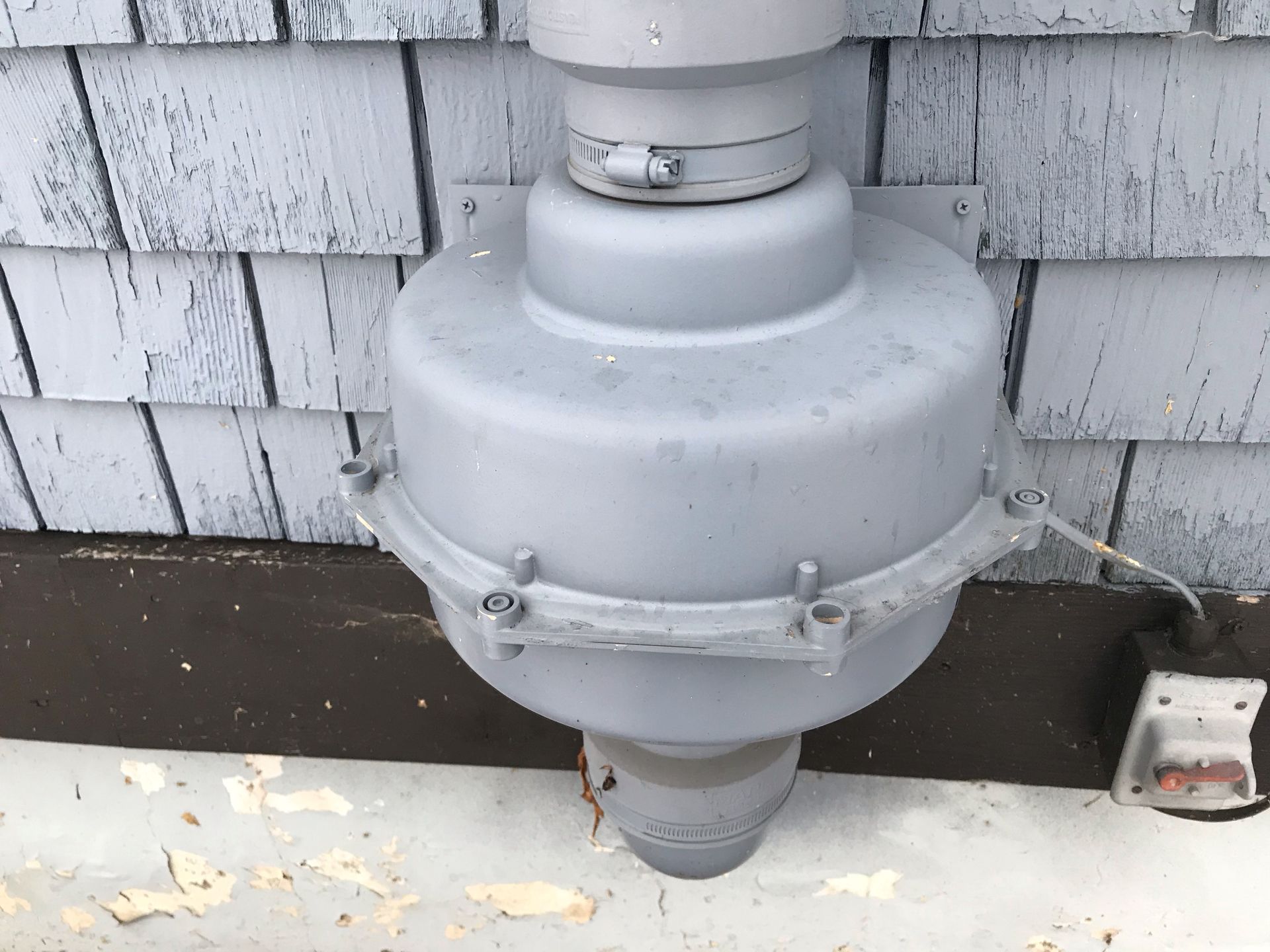 Gray rooftop vent pipe against weathered shingles and siding, with a small electrical box nearby