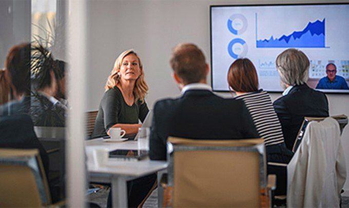 Business meeting with woman speaking, screen with graphs, and person on video call.