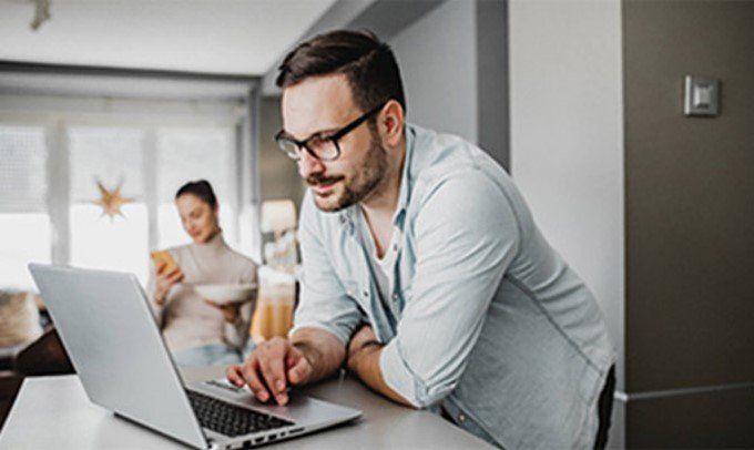 Man with glasses leaning on a counter, typing on a laptop. A woman is blurred in the background, looking at her phone.