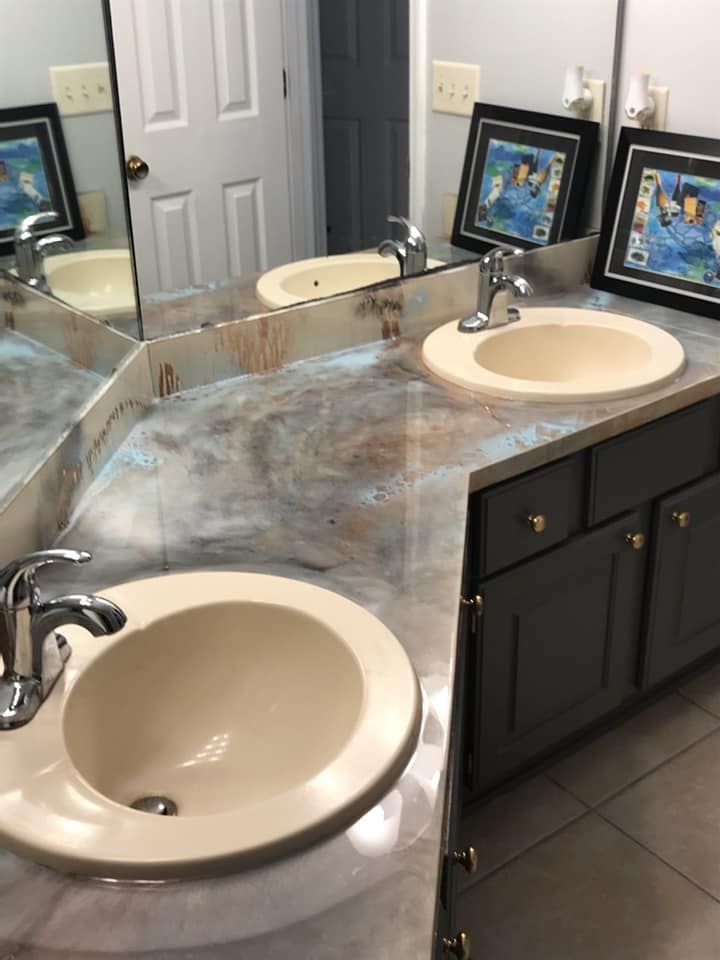 Bathroom with two sinks, gray cabinets, silver faucets, and a speckled countertop. Two framed pictures sit on the counter.