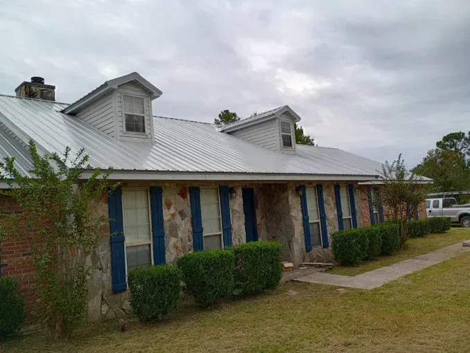 House with gray metal roof, stone facade, blue shutters, and green bushes under an overcast sky.