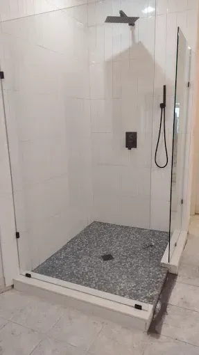 Modern shower with pebble floor, white tiled walls, glass doors, and black fixtures.