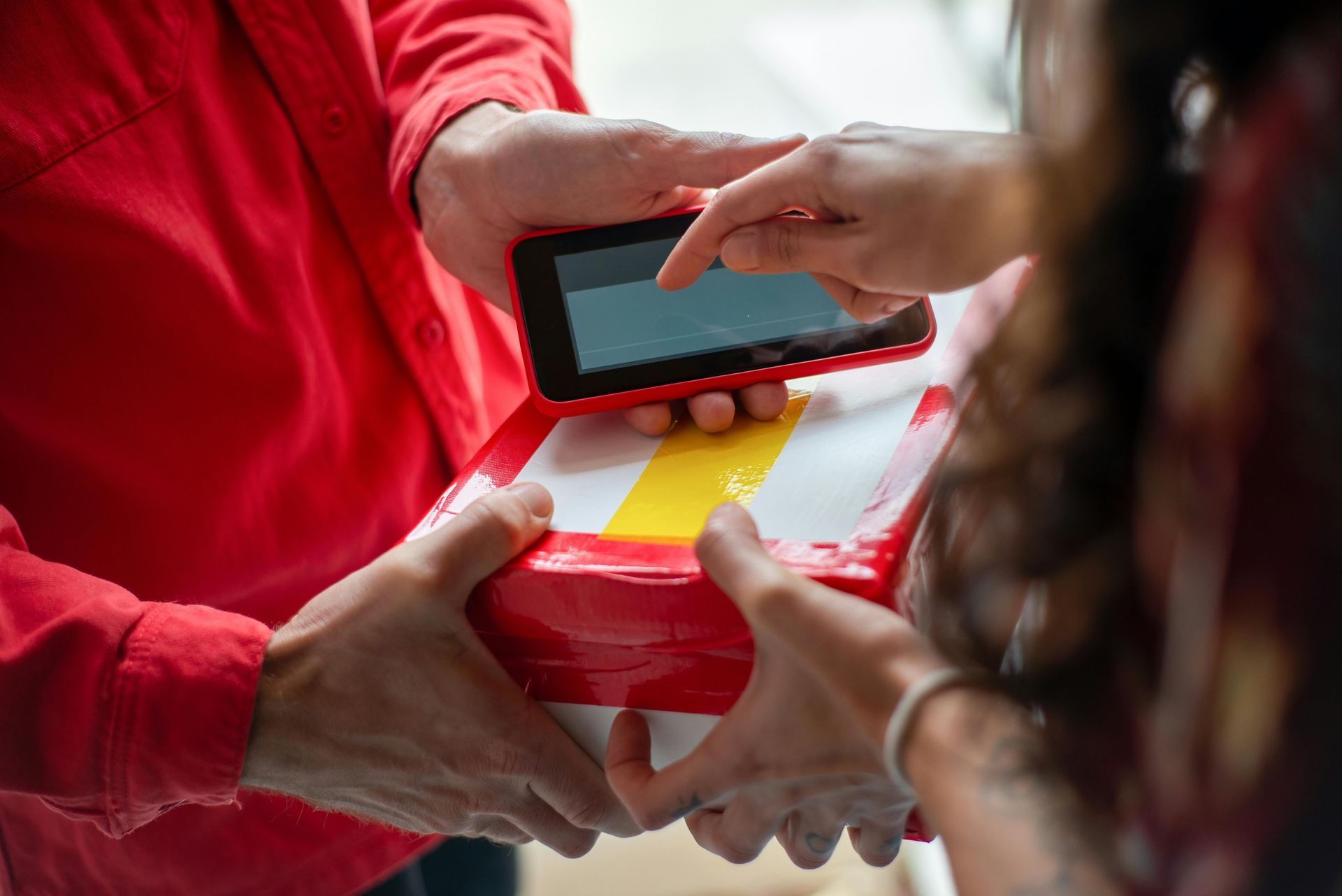 Person in red uniform hands a package to another person, who signs a tablet held on top.