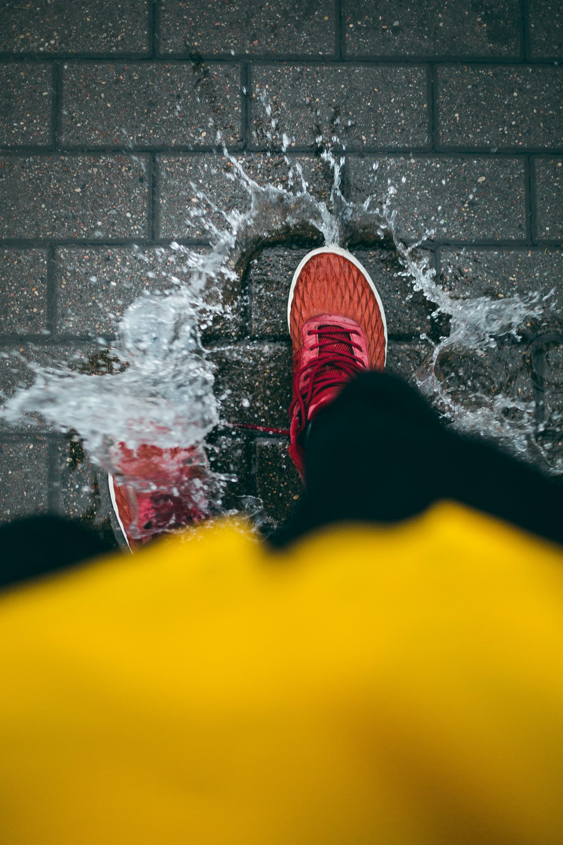 Person stepping in a puddle, water splashing. Red shoe, black pants, yellow coat, wet brick sidewalk.
