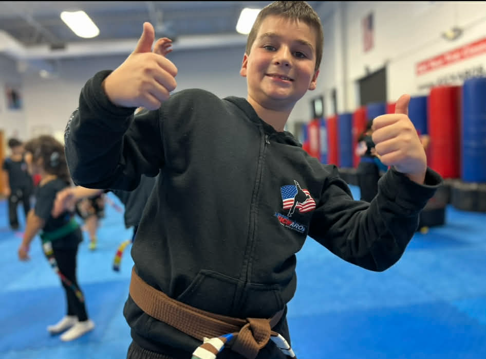 Boy in brown belt and black hoodie gives two thumbs up in a martial arts gym.