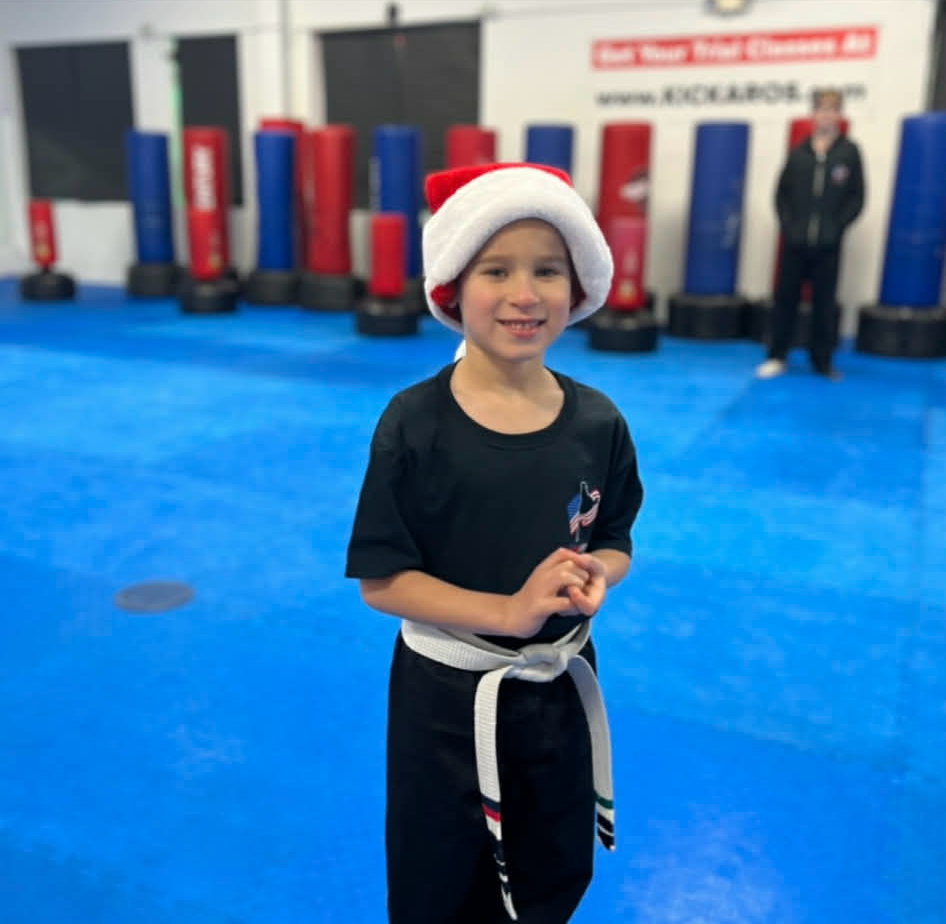 Child in Santa hat, karate uniform, white belt, smiles in a gym, with punching bags and other person in background.