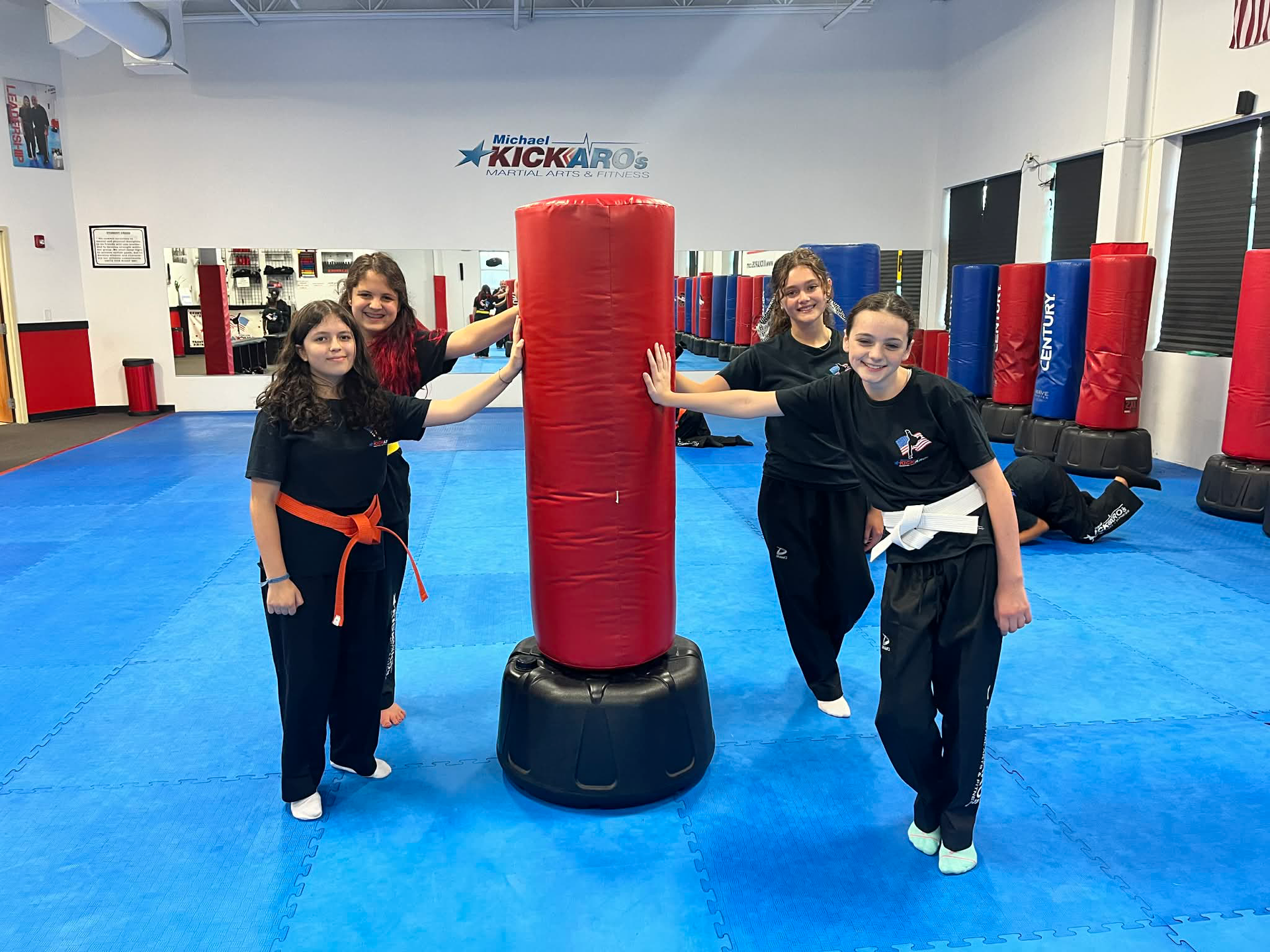 Four martial arts students, wearing black uniforms, pose by a red punching bag in a studio with blue mats.