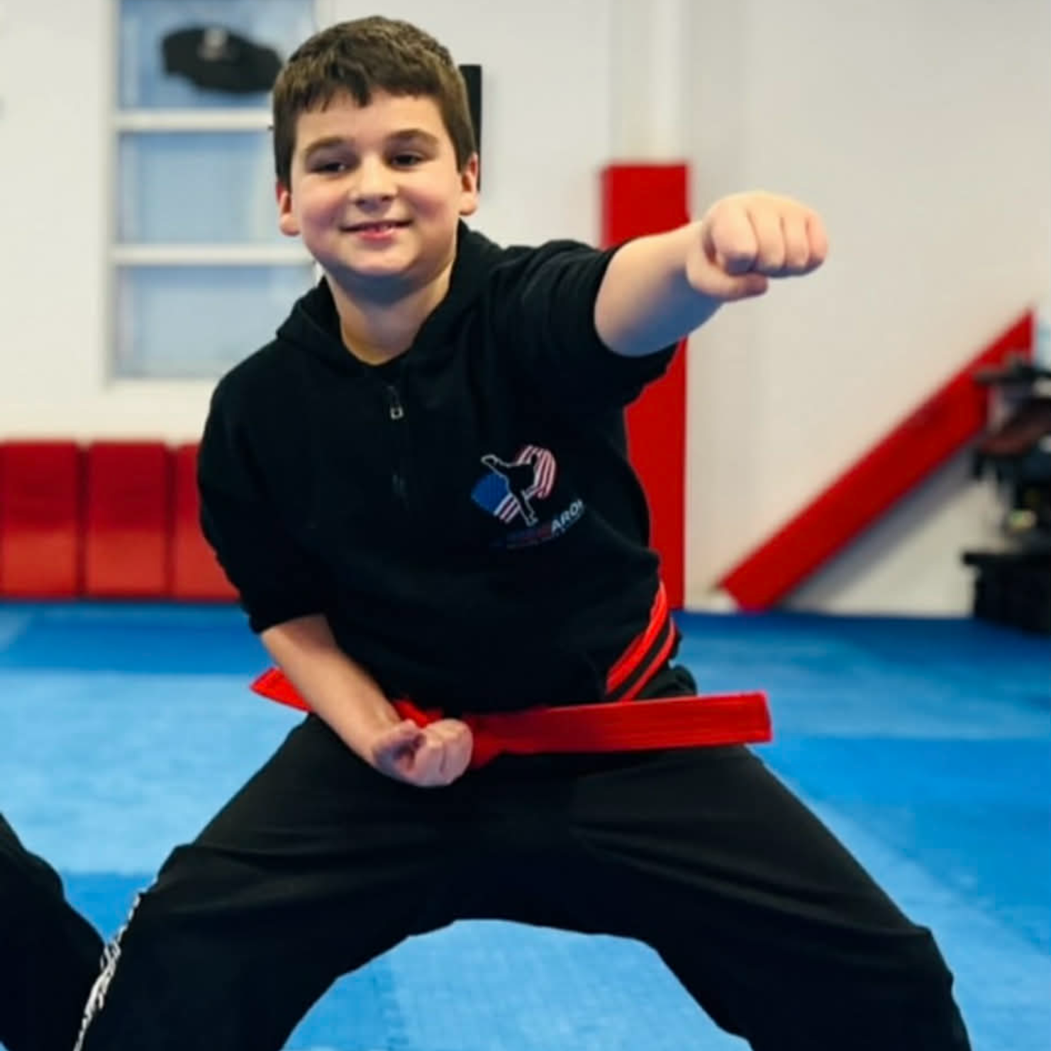 A student in a black martial arts uniform and a red belt performs a forward punch in a gym with blue floor mats.