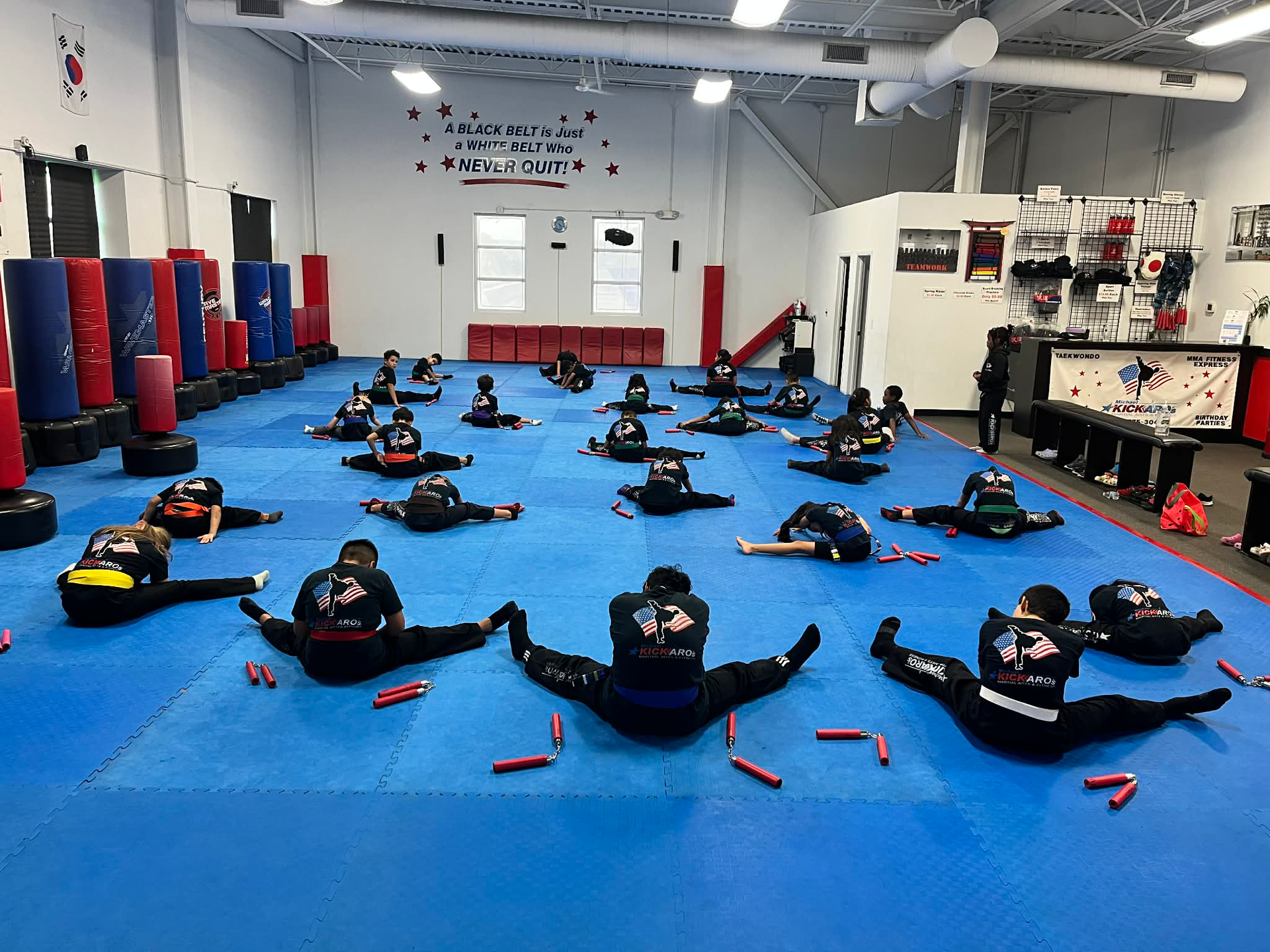 Martial arts students in blue uniforms stretching on blue mats in a gym.