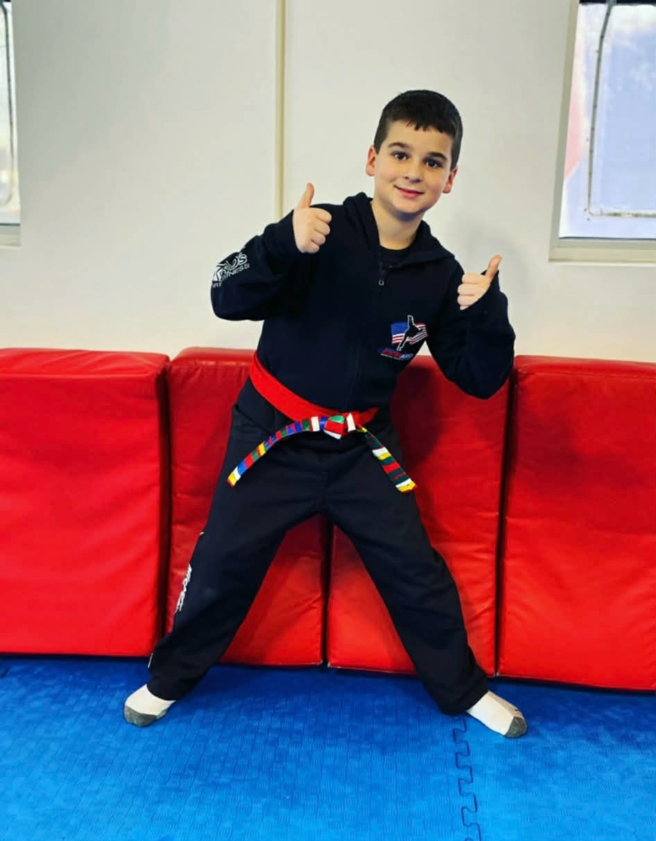 Boy in martial arts uniform with thumbs up. Standing in a dojo with red and blue mats.