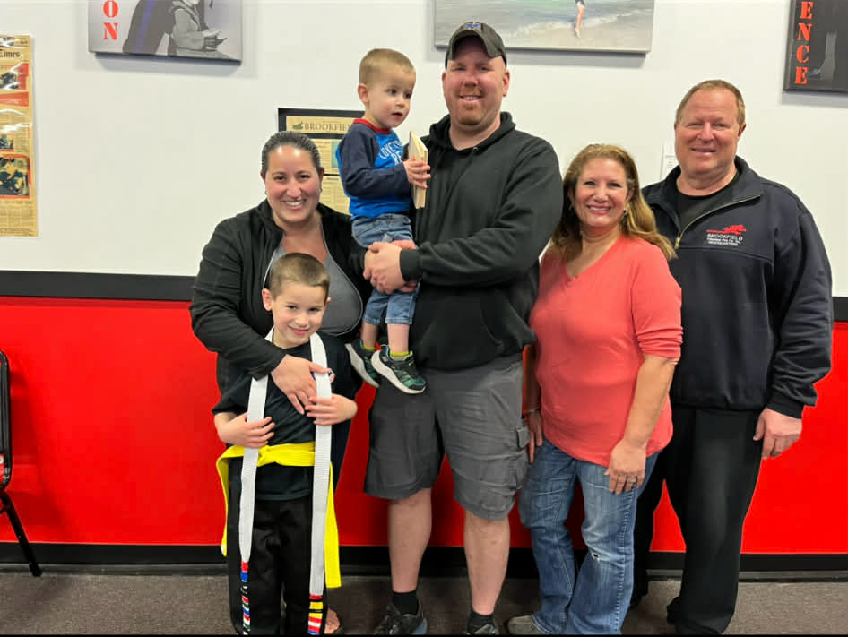 Group poses for photo at martial arts studio. Two adults hold children, others smile. Black, red decor.