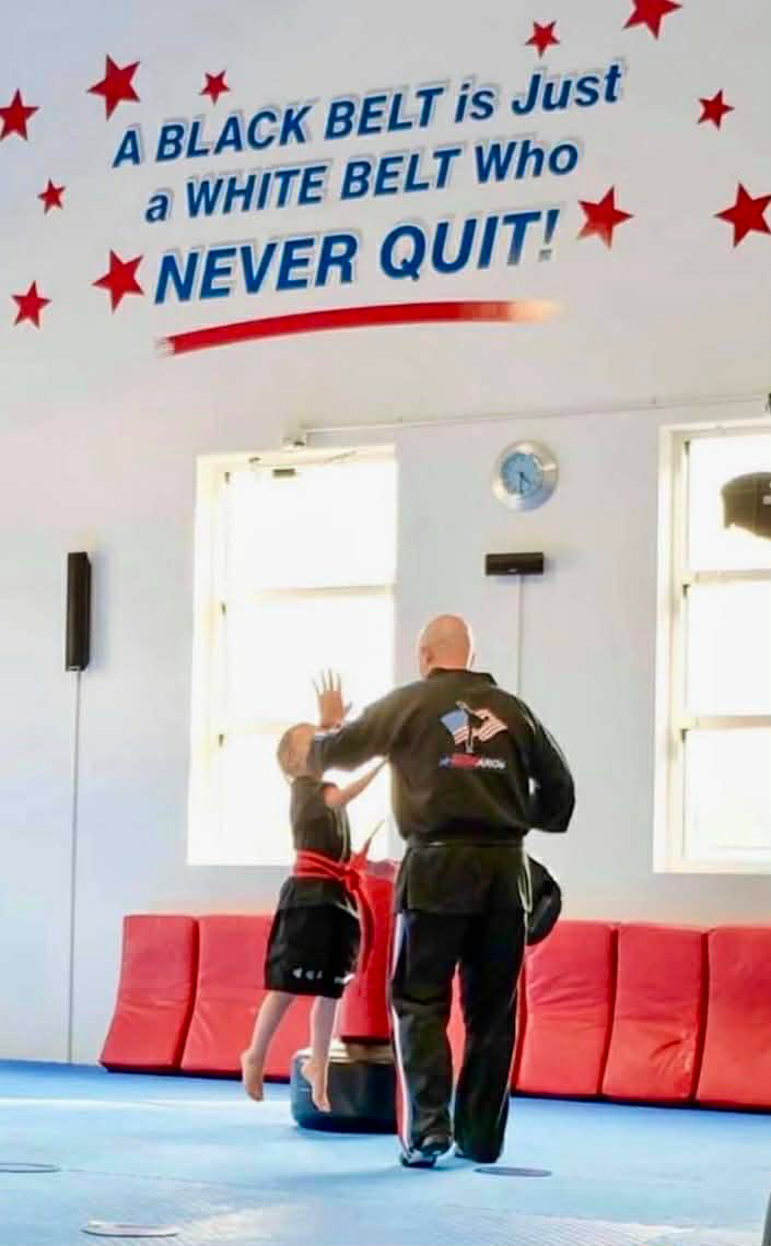 Instructor and student in martial arts training, classroom setting. Text: “A Black Belt is Just a White Belt Who Never Quit!”