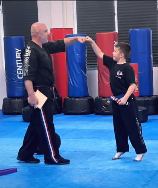 Man in black uniform fist-bumps boy in karate uniform, training room with punching bags in background.