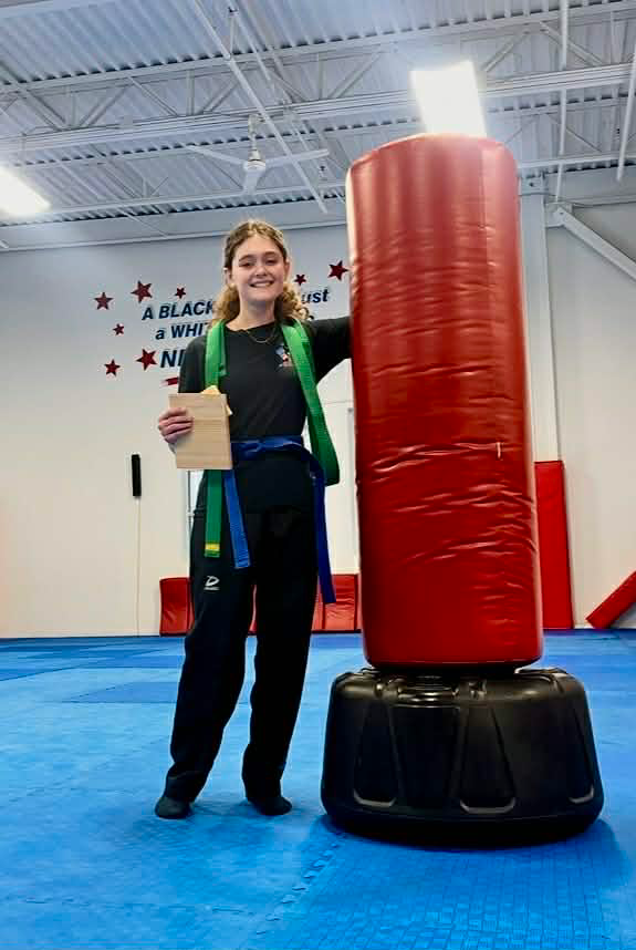 Teenager in martial arts attire stands by a red punching bag, holding a wooden board, in a dojo.
