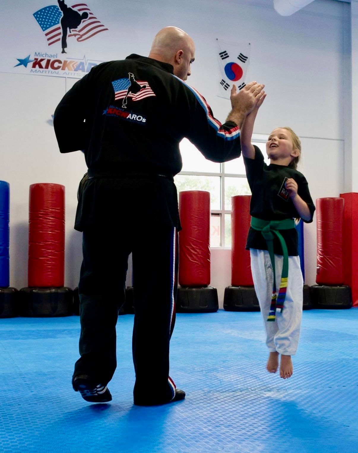 A martial arts instructor stands on a blue mat, giving a high-five to a leaping student in a black uniform and green belt.