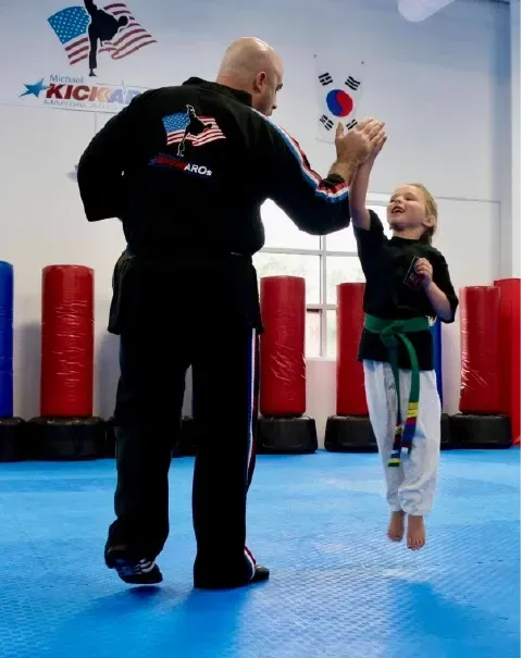 A martial arts instructor gives a high-five to a leaping student on a blue mat in a gym with punching bags.