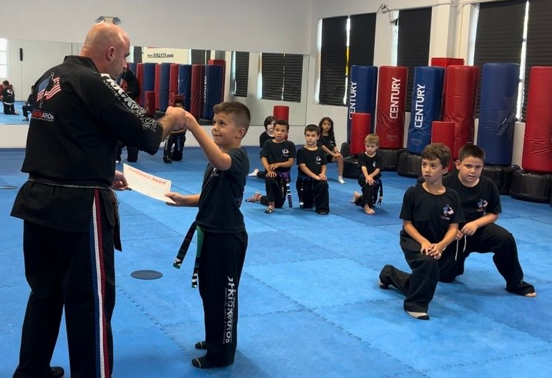 An instructor in a black uniform hands a certificate to a student in a martial arts studio with other students watching.