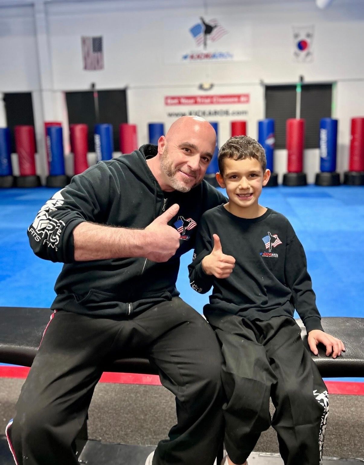 Two people seated on a gym mat, smiling and giving thumbs-up in a martial arts training studio