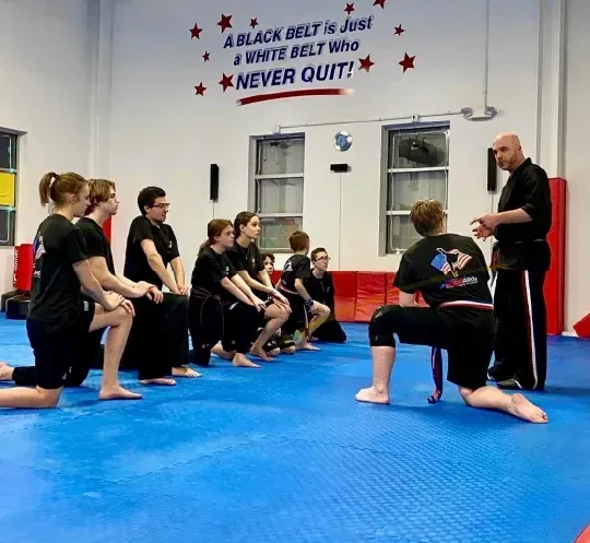 A group kneels on a blue mat in a martial arts studio while a teacher leads a lesson under an inspirational sign.