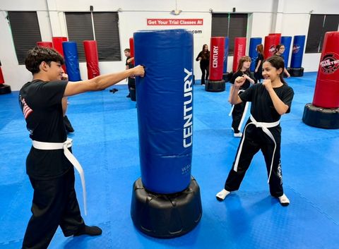 Students in black uniforms practice boxing on tall blue and red Century punching bags in a bright martial arts studio.