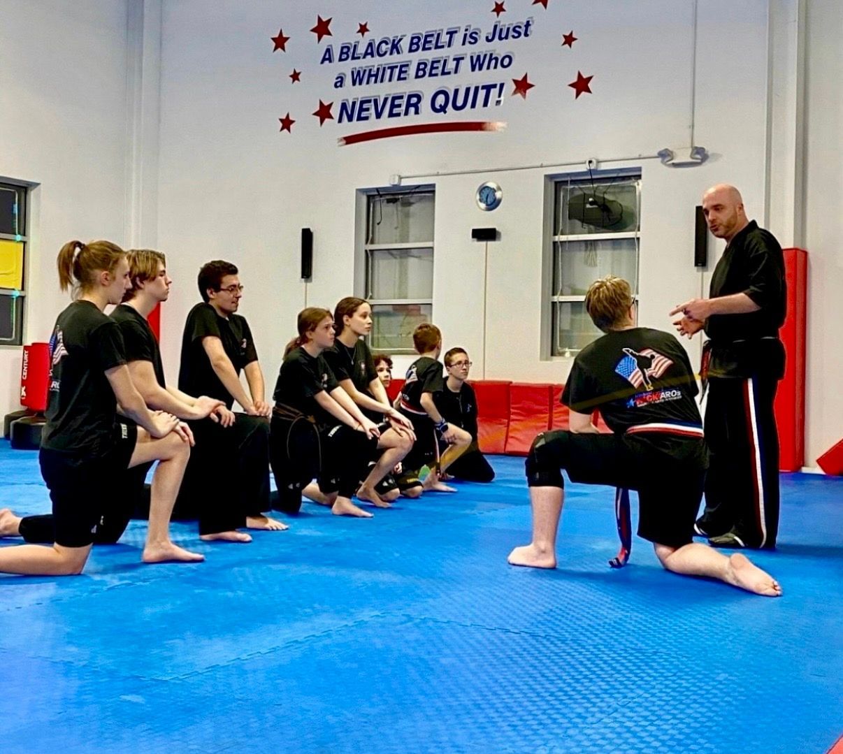 A group kneels on a blue mat in a martial arts studio while a teacher leads a lesson under an inspirational sign.