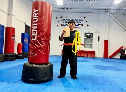 A martial artist stands next to a red Century punching bag holding a wooden board, wearing a uniform and a yellow belt.