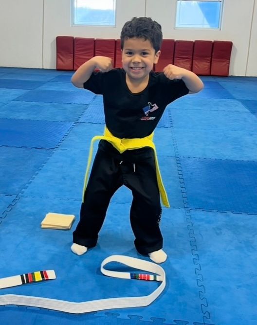 A person wearing a martial arts uniform with a yellow belt flexes their muscles on a blue matted floor.