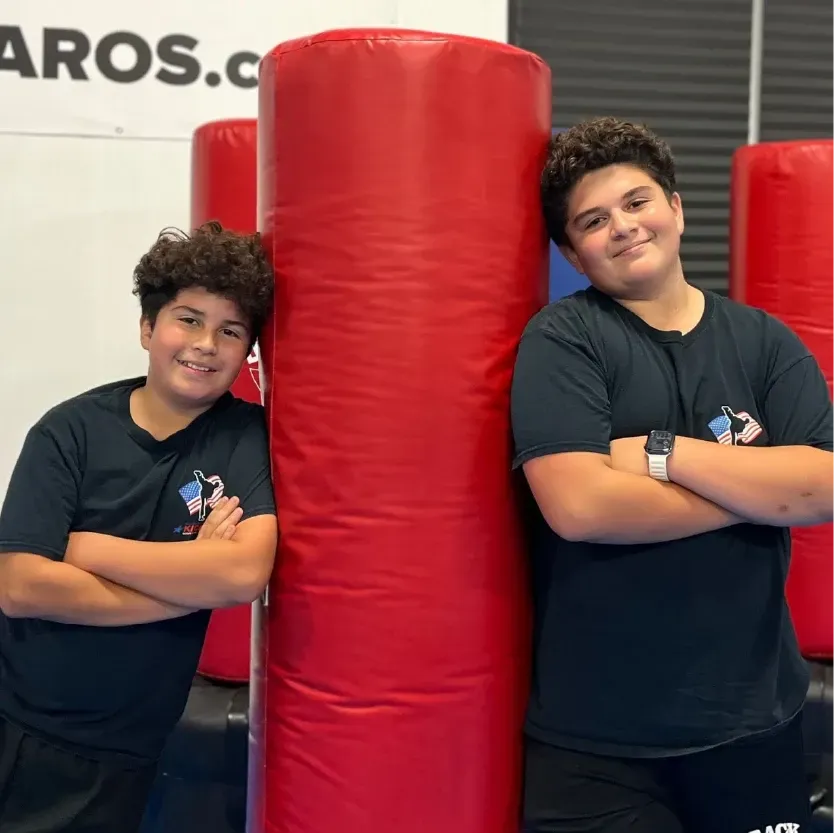 Two people in dark shirts stand with arms crossed on either side of a large red cylindrical exercise bag in a gym.