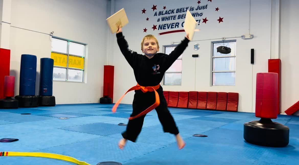 Young martial artist, in black uniform, jumps with joy, holding broken boards. Blue mat, training space.