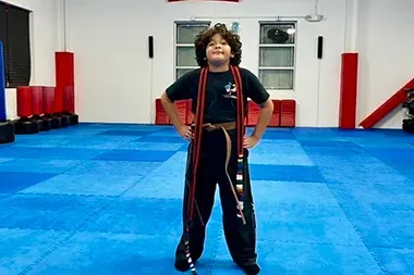 Boy in martial arts uniform stands with jump rope in gym, hands on hips. Blue mats, motivational quote.